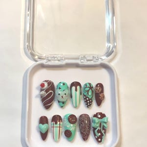 Puede incluir: Un juego de diez uñas postizas en forma de almendra en una caja de plástico transparente. Las uñas presentan varios diseños, incluyendo goteos de chocolate, una cara de oso, acentos verde menta y un lazo. La paleta de colores incluye marrón, blanco y verde menta.