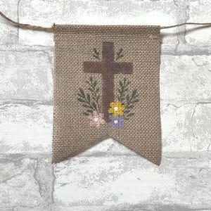 May include: A burlap banner with a brown cross, green leaves, and colorful flowers. The banner is suspended by a natural twine string, perfect for Easter or religious decor.