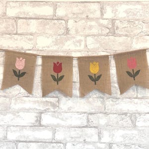 May include: A burlap banner with four pennants, each featuring a tulip design. The tulips are in pink, red, yellow, and pink, with green stems and leaves. The banner is strung on a thin brown cord, perfect for spring decor.