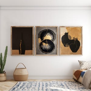 May include: Three framed wall art prints featuring abstract depictions of pottery with gold-coloured cracks. The left print shows a black vase silhouette, the centre shows two circular black forms, and the right shows two black vessels on a tan background.