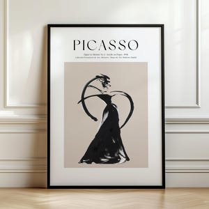 May include: A framed vertical art print featuring the text PICASSO at the top, followed by smaller text. The central image is a black abstract ink-style figure on a beige background, depicting a person in a flowing dress with curved lines.