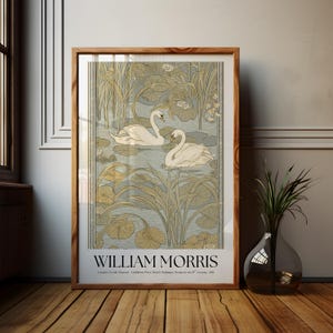 May include: A framed art print featuring two white swans swimming in a pond surrounded by water lilies and tall reeds. The print includes the text WILLIAM MORRIS and London Textile Museum Exhibition Prints British Wallpaper Design in the 19th Century 2011.