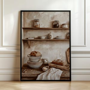 May include: A framed wall art print depicting a rustic kitchen scene with wooden shelves holding glass jars and ceramic bowls. A stack of plates topped with a loaf of bread sits on a wooden counter next to a cutting board, a small dish, and a white cloth.