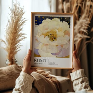 May include: A framed art print featuring a large, pale yellow and white flower with a dark blue background. The text at the bottom reads KLIMT 1862-1918, ROMA 1891, VER SACRUM, WIENER SECESSION. The frame is light wood and rectangular in shape.