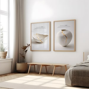 May include: Two framed wall art prints featuring white ceramic vessels repaired with gold lacquer in the Kintsukuroi style. Each print includes the text Kintsukuroi and a description of the art form, displayed in light wood frames on a white wall.