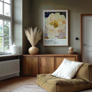 May include: A framed art print featuring a large white peony flower with yellow centers and a dark blue background, labeled KLIMT 1862-1918. The print is displayed on a wall above a wooden cabinet, next to a vase of pampas grass and a low-profile chair.