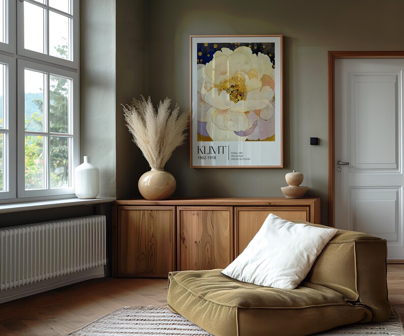May include: A framed art print featuring a large white peony flower with yellow centers and a dark blue background, labeled KLIMT 1862-1918. The print is displayed on a wall above a wooden cabinet, next to a vase of pampas grass and a low-profile chair.