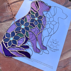 Könnte beinhalten: Ein Glasmalerei-Hunde-Silhouette mit floralen Details in Lila-, Blau- und Grüntönen. Das Kunstwerk ist mit Kupfer umrandet und enthält eine Aufhängekette. Ein Papier mit einer Hundeumrisslinie und dem Text "SUNNY PANES STUDIO" befindet sich hinter dem Glas.