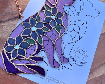 May include: A stained glass dog silhouette with floral details in shades of purple, blue, and green. The artwork is outlined in copper and includes a hanging chain. A paper with a dog outline and text "SUNNY PANES STUDIO" is behind the glass.