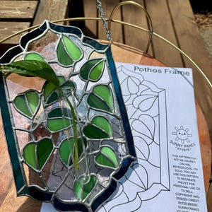 以下が含まれることがあります： 緑色の植物の茎が入った透明なガラスバイアルが付いた、ステンドグラスのポトスフレーム。フレームは、葉のデザインの緑と透明なガラスが特徴で、濃い青色のボーダーが付いています。「Pothos Frame」と「SUNNY PANES STUDIO」の文字が見えます。