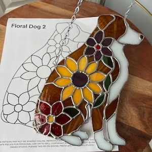 Könnte beinhalten: Glasmalerei-Hund mit floralem Design. Der Hund ist braun, weiß und gelb, mit einer Sonnenblume und anderen Blumen. Der Text "Floral Dog 2" ist auf einem Papier im Hintergrund zu sehen. Er hängt an einer silbernen Kette.