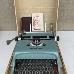 Può includere: Una macchina da scrivere Underwood-Olivetti turchese vintage nella sua custodia da trasporto beige originale. La macchina da scrivere ha tasti neri con lettere bianche e un tasto di ritorno rosso. La custodia è aperta, rivelando la macchina da scrivere, un libretto e una piccola carta con grafica colorata.