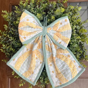 May include: A decorative wreath featuring a large, quilted bow with a daisy pattern in yellow and white, outlined in light green. The bow is centered on a lush green wreath, creating a cheerful, spring-like aesthetic.