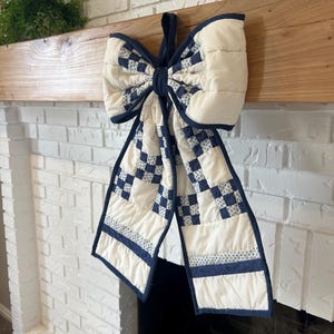 May include: A large, quilted bow decoration in cream and navy blue hangs from a wooden beam. The bow features a checkerboard pattern and solid navy blue trim. The background includes a white brick fireplace and greenery.