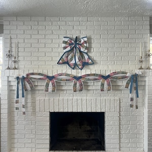 Può includere: Un camino in mattoni bianchi è decorato con un fiocco e una ghirlanda trapuntati. Il fiocco è centrato sopra il camino e la ghirlanda è appesa sopra il caminetto. La ghirlanda e il fiocco presentano motivi floreali con accenti blu e rossi.