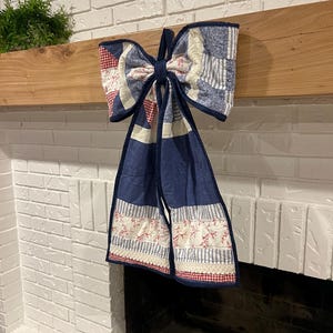 May include: A large, decorative bow made of quilted fabric, featuring a patchwork design in shades of blue, red, and white. The bow is hanging from a wooden beam, with long, flowing tails. The bow is likely used as a holiday or seasonal decoration.