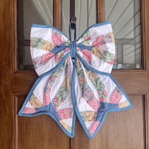 May include: A large, quilted bow decoration with a blue border and a white centre. The bow features sections of floral and patterned fabric in pink, green, and yellow. It hangs from a brown door hanger on a wooden door.