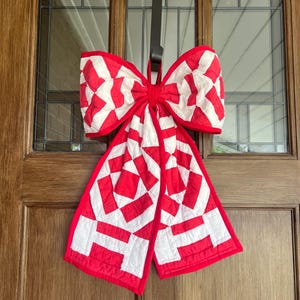 May include: A large, quilted bow in red and white hangs on a wooden door. The bow features a geometric pattern of red and white squares and rectangles. The bow is a decorative item, possibly for a holiday or special occasion.