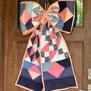May include: A large decorative bow crafted from quilted fabric featuring a patchwork design in shades of navy blue, dusty blue, coral pink, and white. The bow has two long hanging tails and is finished with a solid coral pink fabric trim.