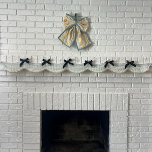 May include: A decorative bow and valance set against a white brick fireplace. The bow features yellow and white fabric with a green trim. The valance is light blue with black bow accents and gold trim, adding a touch of elegance to the fireplace.