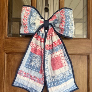 May include: A large, quilted bow door decoration in shades of red, white, and blue. The bow features a patchwork design with floral patterns and a dark blue border. It is hanging on a wooden door with a brown strap.
