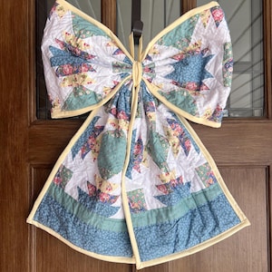 May include: A quilted bow-shaped door decoration. The bow features a patchwork design with white, blue, and floral patterned fabrics. The edges are trimmed with yellow binding. It hangs from a brown strap.