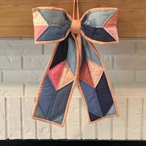 May include: A quilted fabric bow featuring a geometric patchwork design in shades of navy blue, light blue, dusty pink, and cream. The bow is finished with a peach-colored fabric binding along the edges and includes a matching fabric hanging loop.
