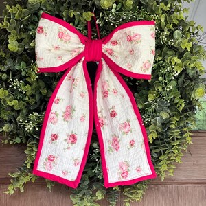 May include: A large decorative bow made of quilted fabric featuring a cream background with a pink rose floral pattern. The bow has a bright pink fabric border along all edges and a central knot, designed to be hung as a wall or door ornament.