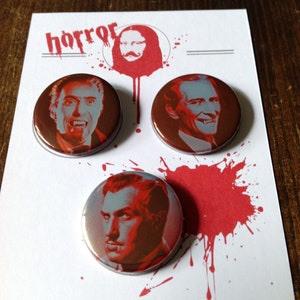 May include: Three round, silver-coloured buttons with red accents. Each button features a black and white image of a man's face. The buttons are arranged in a triangular pattern on a white background with red splatter paint. The word "horror" is printed in black at the top of the background.