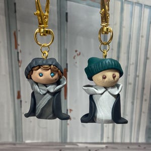 May include: Two handmade keychains with gold-colored clasps. Each keychain features a small, detailed figurine. One figurine wears a dark gray hooded cloak and the other wears a green beanie and a white collar. Both have gold-colored eyes.