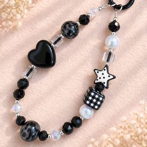 May include: A black beaded keychain with a variety of decorative beads. The keychain features a black heart, a star with white polka dots, and a checkered cube. Other beads include black, iridescent, and speckled gray spheres.