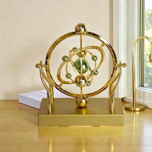 Può includere: Un ornamento da scrivania dorato che rappresenta un modello del sistema solare. Presenta una sfera verde centrale circondata da anelli orbitali e sfere verdi più piccole. Poggia su una base rettangolare dorata.
