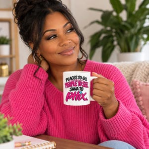 Pode incluir: Uma caneca de cerâmica branca com texto em preto e rosa que diz Places to go. People to see. Java to Phix. com um pequeno ícone de coração azul claro acima da palavra Phix. A caneca tem uma alça padrão e um acabamento liso e brilhante.
