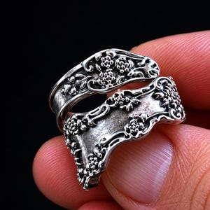 Può includere: Un anello a cucchiaio argentato con dettagli floreali e a volute. L'anello si avvolge attorno al dito, mostrando l'intricato design. L'anello è tenuto su uno sfondo scuro.