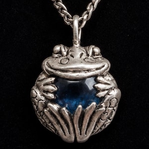 Puede incluir: Un collar con colgante de rana plateada con una gema azul en el centro. La rana tiene detalles y está suspendida de una cadena plateada. El fondo es negro.