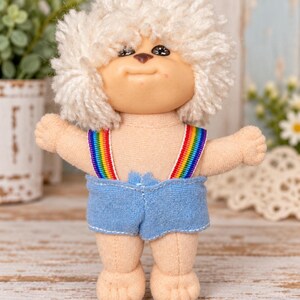May include: A small fabric doll with a round head, tan skin, and thick, curly white yarn hair. The doll features a small brown nose and wears light blue fabric shorts held up by thin, vibrant rainbow-striped suspender straps.