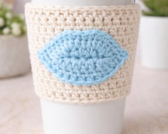 May include: A white ceramic travel mug with a white lid, wrapped in a cream-colored crocheted cozy. The cozy features a light blue crocheted lip design. The mug is on a light pink surface.