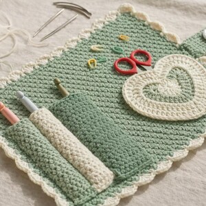 May include: A sage green crochet organizer case with a scalloped cream border, featuring three pockets holding crochet hooks, a heart-shaped applique, small red scissors, and colorful stitch markers, secured by a button tab closure.