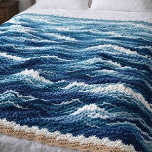 Może przedstawiać: Szydełkowy koc z wzorem fal w odcieniach niebieskiego i białego, przypominający fale oceanu. Koc ma beżową obwódkę, tworząc efekt plaży. Koc jest prezentowany na łóżku z białymi prześcieradłami.