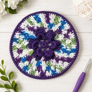 May include: A handmade, circular crochet doily featuring a central flower design in deep purple. The doily has a vibrant color palette, including white, green, blue, and purple. The outer edge is a solid purple, and a crochet hook is visible.