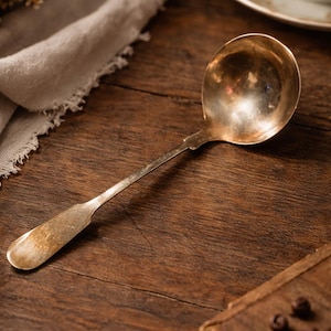 May include: A vintage silver ladle with a long handle and a rounded bowl, resting on a dark wooden surface. The ladle has a tarnished appearance, suggesting age and use. The scene evokes a sense of nostalgia and classic kitchenware.