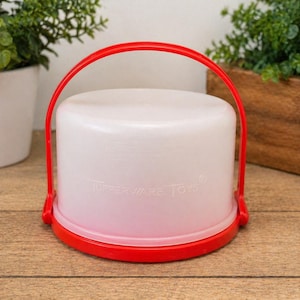 Könnte beinhalten: Ein Vintage-Behälter von Tupperware Toys. Der Behälter ist weiß mit rotem Boden und Griff. Der Griff wölbt sich über die Oberseite. Die Worte "Tupperware Toys" sind auf der Seite aufgedruckt. Der Behälter ist rund und hat einen Deckel.