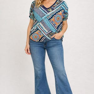 May include: A woman wearing a patterned V-neck top and blue jeans. The top features a patchwork design in shades of blue, orange, and white. The jeans are flared at the bottom, and the woman is wearing brown boots.