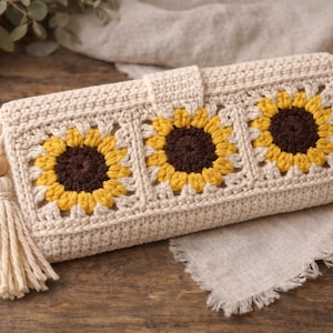 May include: A cream-coloured crochet wallet featuring three sunflower designs with yellow petals and brown centres. The wallet has a tassel and wooden bead detail. The crochet pattern is visible, and the wallet is closed with a flap.