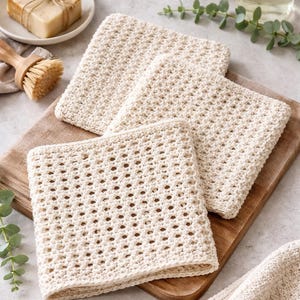 May include: Off-white, square crocheted washcloths with an open weave pattern are displayed on a wooden cutting board. A bar of soap, a wooden brush, and a bottle of liquid soap are also visible.