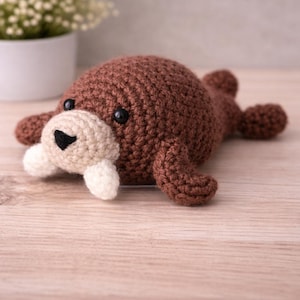 May include: A handmade brown crochet walrus plush toy with a beige snout and white tusks. The stuffed animal has black safety eyes and is lying on a light wood surface. The walrus has small flippers and a round body.