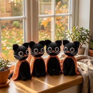 May include: Four handmade black cat plushies with orange capes sit on a wooden windowsill. Each cat has large white eyes, a white mustache, and black ears. The background shows a window with a view of fall foliage and potted plants.