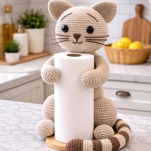 May include: Crocheted cat-shaped paper towel holder in beige and brown. The cat has large black eyes, whiskers, and holds a roll of white paper towels. The holder sits on a wooden base, with a striped tail.