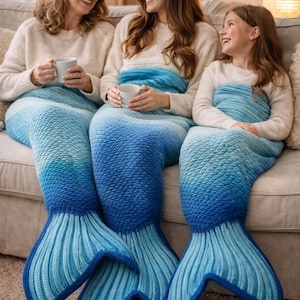 May include: Three people are seated on a couch, each wrapped in a blue ombre mermaid tail blanket. The blankets transition from light blue to dark blue, with a detailed fin design. They are holding white mugs and smiling.