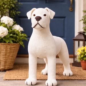 Op de afbeelding: Een groot, handgemaakt, wit gehaakt hondenbeeld staat op een bruine mat. De hond heeft bruine ogen, een bruine neus en zwarte accenten op zijn poten. De achtergrond omvat een blauwe deur, planten en een krans.